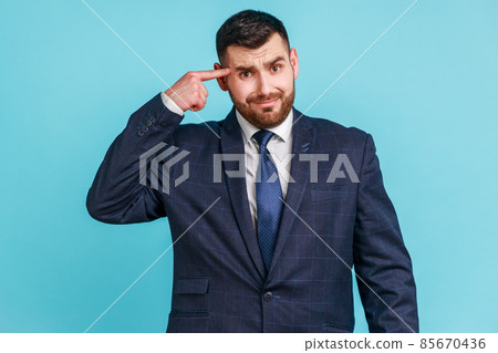 Crazy idea. Portrait of man in dark suit showing stupid gesture, looking at camera with condemnation and blaming for insane plan, dumb suggestion. Indoor studio shot isolated on blue background. 85670436
