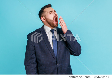 I need rest. Portrait of sleepy man with beard wearing official style suit yawning and covering mouth with hand, feeling exhausted, lack of sleep. Indoor studio shot isolated on blue background. I need rest. Portrait of sleepy man with beard wearing official style suit yawning and covering mouth with hand, feeling exhausted, lack of sleep. Indoor studio shot isolated on blue background. 85670437
