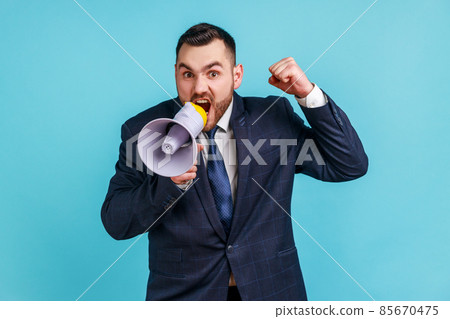 Angry serious businessman wearing dark official style suit loudly speaking screaming holding megaphone, announcing important message. Indoor studio shot isolated on blue background. Angry serious businessman wearing dark official style suit loudly speaking screaming holding megaphone, announcing important message. Indoor studio shot isolated on blue background. 85670475