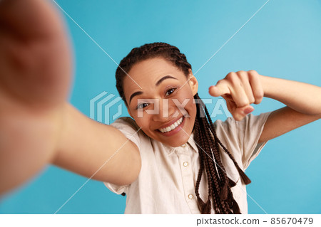 Cheerful woman with black dreadlocks has satisfied face expression, making selfie, expresses sincere emotions, pointing finger to camera, POV. Indoor studio shot isolated on blue background. Cheerful woman with black dreadlocks has satisfied face expression, making selfie, expresses sincere emotions, pointing finger to camera, POV. Indoor studio shot isolated on blue background. 85670479
