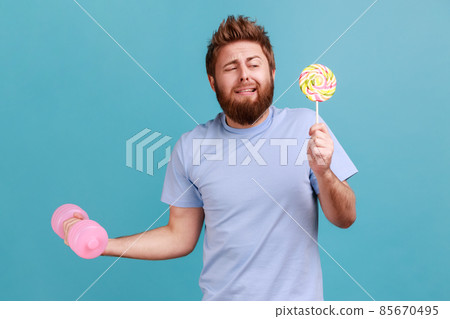 Portrait of funny handsome bearded man holding delicious lollypop and and raised up hand with pink heavy dumbbell, hard to do exercises. Indoor studio shot isolated on blue background. Portrait of funny handsome bearded man holding delicious lollypop and and raised up hand with pink heavy dumbbell, hard to do exercises. Indoor studio shot isolated on blue background. 85670495