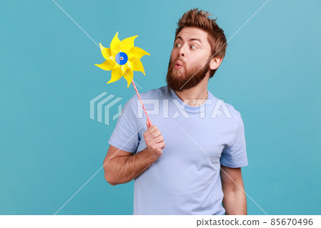 Portrait of funny positive childish handsome bearded man wearing T-shirt playing blowing yellow windmill, having fun with paper toy. Indoor studio shot isolated on blue background. 85670496
