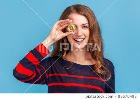 Funny positive woman wearing striped casual style sweater, covering eye with gold bitcoin, having positive expression, crypto currency. Indoor studio shot isolated on blue background. 85670498