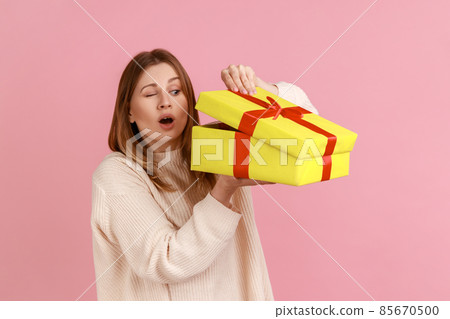 Portrait of curious attractive young blond woman looking inside yellow present box, being interested what inside, wearing white sweater. Indoor studio shot isolated on pink background. Portrait of curious attractive young blond woman looking inside yellow present box, being interested what inside, wearing white sweater. Indoor studio shot isolated on pink background. 85670500