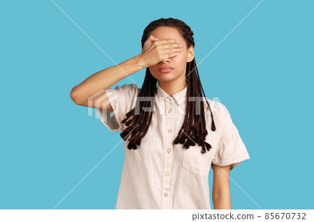 Young woman with black dreadlocks covering her eyes with palm, tries to hide from everybody, dontwant see someone face, wearing white shirt. Indoor studio shot isolated on blue background. 85670732