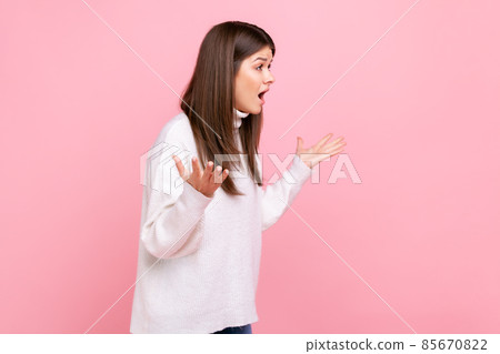 Side view portrait of young girl holding hands in mad furious gesture, screaming with rage and anger, wearing white casual style sweater. Indoor studio shot isolated on pink background. Side view portrait of young girl holding hands in mad furious gesture, screaming with rage and anger, wearing white casual style sweater. Indoor studio shot isolated on pink background. 85670822