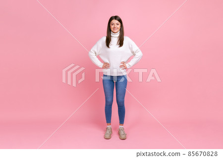 Full length portrait of beautiful brunette female standing with hands on hips, looking at camera, wearing white casual style sweater. Indoor studio shot isolated on pink background. Full length portrait of beautiful brunette female standing with hands on hips, looking at camera, wearing white casual style sweater. Indoor studio shot isolated on pink background. 85670828