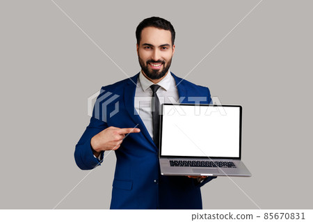 Optimistic man pointing finger at laptop with blank display, showing empty screen, bragging with internet advertisement, wearing official style suit. Indoor studio shot isolated on gray background. Optimistic man pointing finger at laptop with blank display, showing empty screen, bragging with internet advertisement, wearing official style suit. Indoor studio shot isolated on gray background. 85670831
