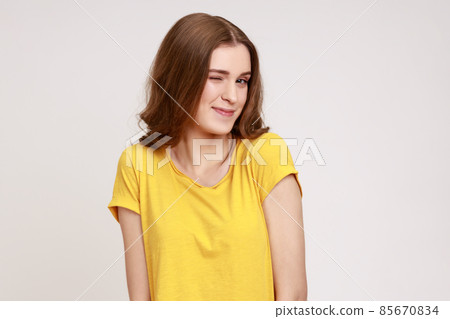 Closeup portrait of playful happy charming teenager girl with wavy hair in casual yellow T-shirt blinking eye, having cunning idea, flirting to camera. Indoor studio shot isolated on gray background. 85670834