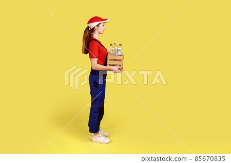 Side view portrait of positive satisfied woman volunteer standing with box full of empty plastic bottles, wearing overalls and red cap. Indoor studio shot isolated on yellow background. Side view portrait of positive satisfied woman volunteer standing with box full of empty plastic bottles, wearing overalls and red cap. Indoor studio shot isolated on yellow background. 85670835