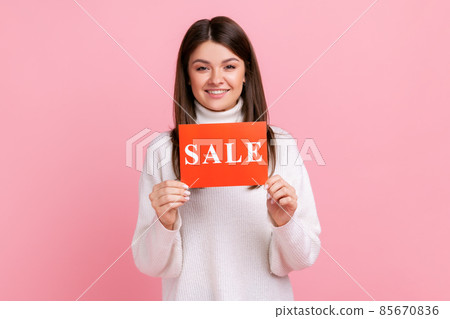 Positive brunette female holding card with sale inscription, looking at camera with toothy smile, wearing white casual style sweater. Indoor studio shot isolated on pink background. Positive brunette female holding card with sale inscription, looking at camera with toothy smile, wearing white casual style sweater. Indoor studio shot isolated on pink background. 85670836
