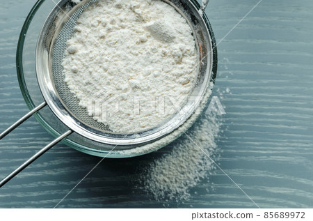Wheat flour is sown through a sieve into a glass bowl through a metal sieve, on a wooden table. Wheat flour is sown through a sieve into a glass bowl through a metal sieve, on a wooden table. 85689972