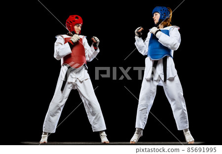 Dynamic portrait of two young women, taekwondo athletes training together isolated over dark background. Concept of sport, skills 85691995