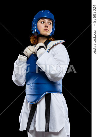 Portrait of young girl, taekwondo practitioner posing isolated over dark background. Concept of sport, skills Portrait of young girl, taekwondo practitioner posing isolated over dark background. Concept of sport, skills 85692024