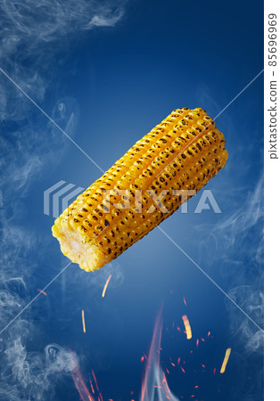 Grilled ear of corn hovering over flames in sparks and haze on blue background Grilled ear of corn hovering over flames in sparks and haze on blue background 85696969