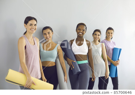 Group portrait of happy diverse girls after yoga workout. Five multiracial young women in modern sports outfits standing by gym wall, holding fitness rubber mats, looking at camera and smiling 85700646