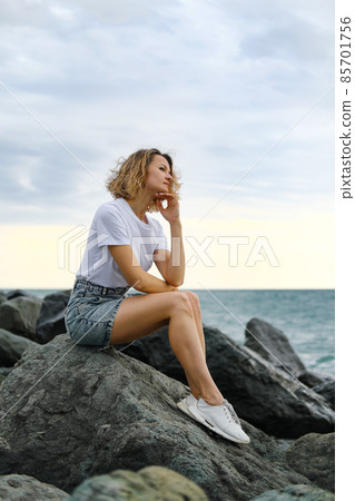 A young attractive Caucasian blonde woman of 35 years sits on the rocks of sea coast. Emerald-colored water, waves splashing on the stones. The woman meditates and relaxes. A young attractive Caucasian blonde woman of 35 years sits on the rocks of sea coast. Emerald-colored water, waves splashing on the stones. The woman meditates and relaxes. 85701756