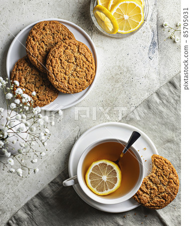 White porcelain cup with lemon tea and oatmeal cookies on textured background with tablecloth and white flowers. Top view 85705031