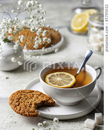 White porcelain cup with lemon tea and oatmeal cookies on textured background with flowers on the back White porcelain cup with lemon tea and oatmeal cookies on textured background with flowers on the back 85705032
