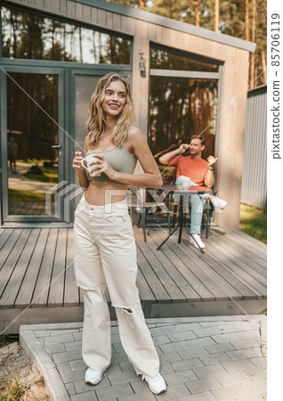 Pretty long-haired blonde woman with a coffee cup standing outsie the cottage Pretty long-haired blonde woman with a coffee cup standing outsie the cottage 85706119