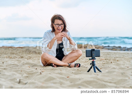 Middle aged woman sitting on beach with smartphone using video call Middle aged woman sitting on beach with smartphone using video call 85706894