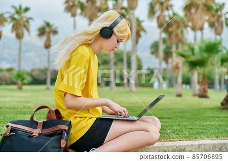 Female teenage student in headphones sitting in park with laptop. Female teenage student in headphones sitting in park with laptop. 85706895