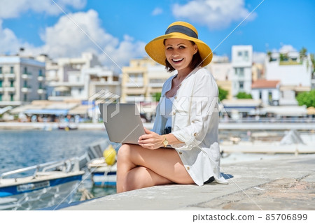 Mature woman in hat with laptop on sea promenade in city Mature woman in hat with laptop on sea promenade in city 85706899