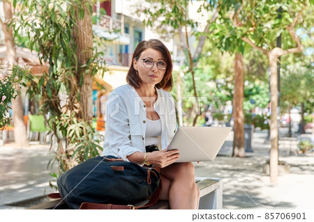 Outdoor portrait of middle-aged business woman sitting on street of city with laptop Outdoor portrait of middle-aged business woman sitting on street of city with laptop 85706901