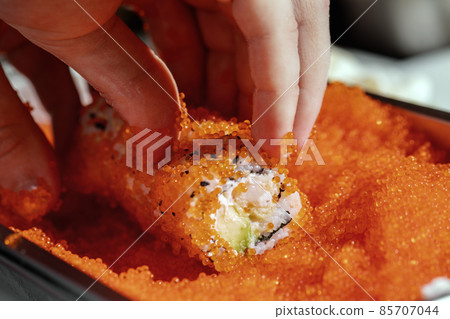 Closeup of chef hands rolling up sushi in flying fish roe (Tobiko) red caviar - focus on the roll. 85707044