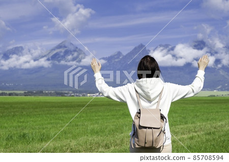 A woman tourist in a white hoodie and a backpack enjoys a beautiful view of the Tatra mountains while standing in a green field. Clouds float over mountain peaks, copy space 85708594