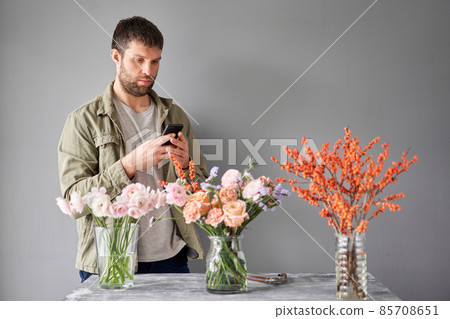 Male florist with a mobile phone, takes a photo to confirm the order. Accepts an online order for flower delivery. Responds to messages in social networks and online messengers. Male florist with a mobile phone, takes a photo to confirm the order. Accepts an online order for flower delivery. Responds to messages in social networks and online messengers. 85708651