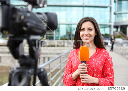 Young female journalist with microphone working on city street Young female journalist with microphone working on city street 85709689