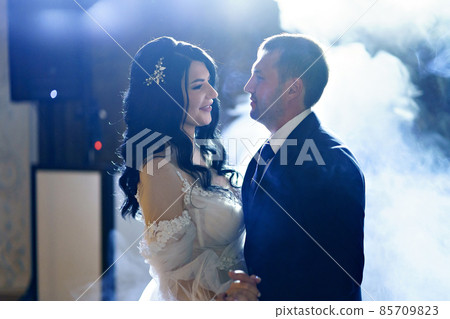 the bride and groom in wedding dresses dance in a dark hall in heavy smoke. 85709823