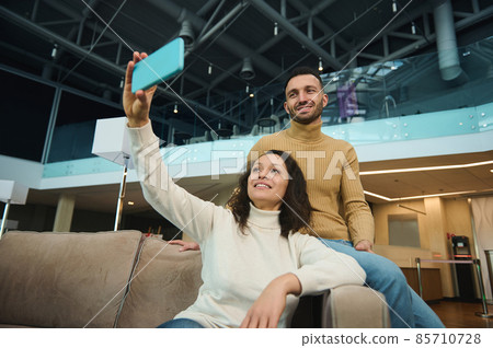 Beautiful young loving couple of newlyweds on their honeymoon trip making selfie on a smartphone while relaxing together in the airport lounge while waiting for boarding a flight. Beautiful young loving couple of newlyweds on their honeymoon trip making selfie on a smartphone while relaxing together in the airport lounge while waiting for boarding a flight. 85710728