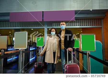 Young couple of business partners in protective medical masks, travel during pandemic, standing with luggage at check-in counter, passing customs and passport control at airport terminal. Copy space 85710749