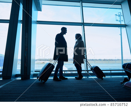 Silhouette of two people, woman and man with suitcases, standing by the panoramic windows overlooking the runways and planes in the departure terminal of the international airport, awaiting a flight 85710752