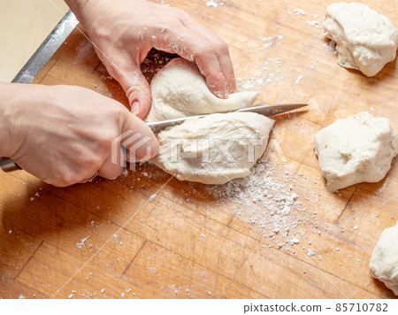 Female hands cut with a knife a kneaded dough on a wooden table. Female hands cut with a knife a kneaded dough on a wooden table. 85710782