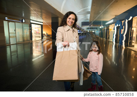 Beautiful woman, happy mother with shopping bags stands with her adorable little daughter in the duty free zone of the departure terminal at the international airport, waiting for the upcoming flight Beautiful woman, happy mother with shopping bags stands with her adorable little daughter in the duty free zone of the departure terminal at the international airport, waiting for the upcoming flight 85710858