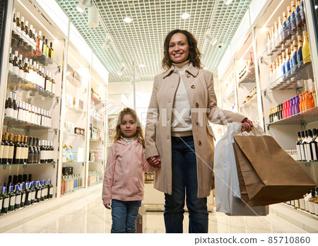 Happy mother and her cute daughter travel together, standing with shopping bags in the duty free store, making purchases while waiting for boarding in the departure terminal of international airport Happy mother and her cute daughter travel together, standing with shopping bags in the duty free store, making purchases while waiting for boarding in the departure terminal of international airport 85710860