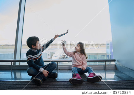 Little boy and girl, brother and sister playing with toy airplanes sitting on the floor by the panoramic windows of the airport departure terminal overlooking the runways. Family air travel concept 85710884