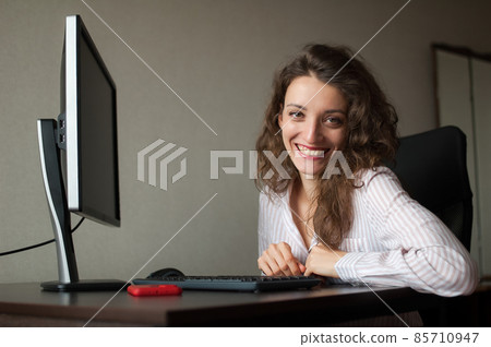 Young female office manager in white shirt and curly hair is sitting at the table and typing using keyboard, routine work, freelance 85710947