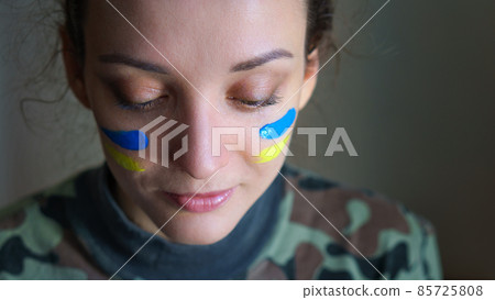 Indoor portrait of young girl with blue and yellow ukrainian flag on her cheek wearing military uniform, mandatory conscription in Ukraine, equality concepts 85725808