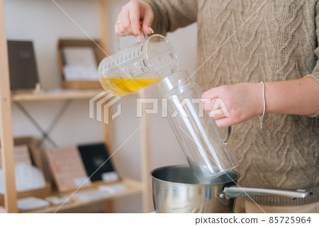 Close-up cropped shot of unrecognizable female artisan pouring melted wax into glass vessel for creating candle building mixture. 85725964