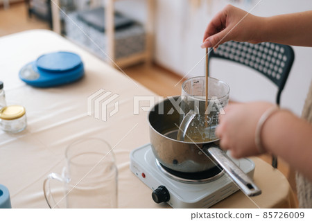 Close-up cropped shot of unrecognizable female artisan melted wax into pot of boiling water for creating candle building mixture. Close-up cropped shot of unrecognizable female artisan melted wax into pot of boiling water for creating candle building mixture. 85726009
