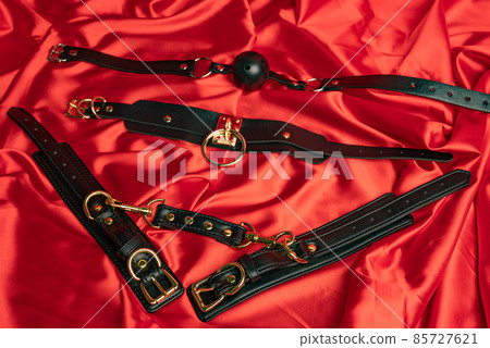 Adult sex games. BDSM items. Leather straps handcuffs, collar and gag ball on a red satin sheet. Adult sex games. BDSM items. Leather straps handcuffs, collar and gag ball on a red satin sheet. 85727621