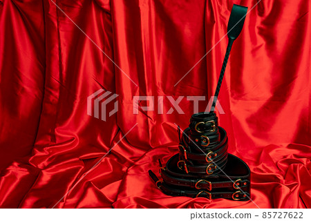 Adult sex games. BDSM items. Leather straps handcuffs, belt and whip on a red satin sheet. Adult sex games. BDSM items. Leather straps handcuffs, belt and whip on a red satin sheet. 85727622