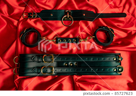 Adult sex games. BDSM items. Leather straps handcuffs, collar on a red satin sheet. Adult sex games. BDSM items. Leather straps handcuffs, collar on a red satin sheet. 85727623