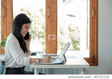 Side view happy asian businesswoman sitting on her workplace in the office. Young woman working for financial document data charts with laptop during call phone in the office Side view happy asian businesswoman sitting on her workplace in the office. Young woman working for financial document data charts with laptop during call phone in the office 85732119