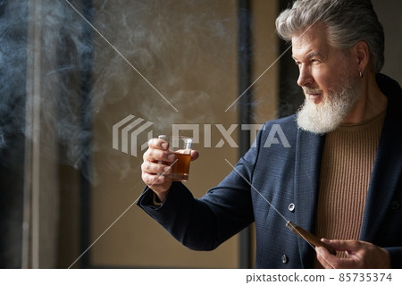 Portrait of sophisticated mature businessman with beard looking away while holding glass of whisky and cigar, standing in loft interior 85735374