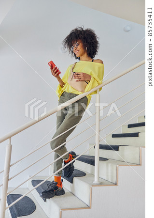 Positive African-American woman in stylish casual clothes looks at mobile phone on staircase 85735411
